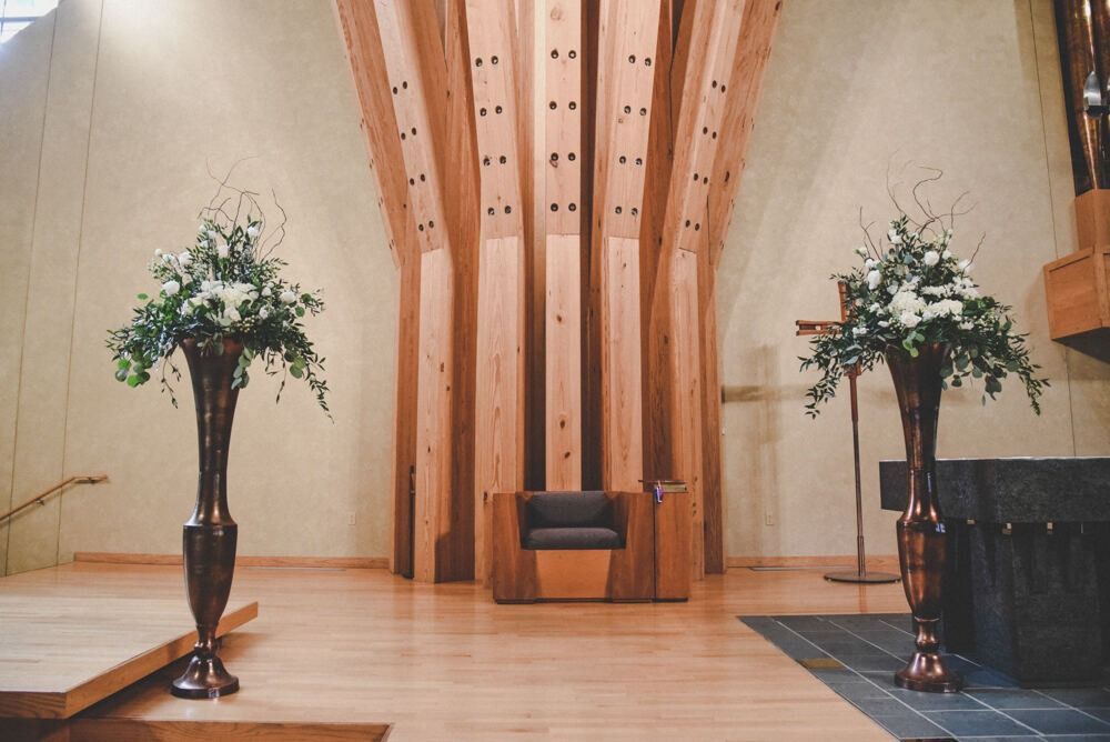 Church interior with two floral arrangements flanking a wooden structure and seating.