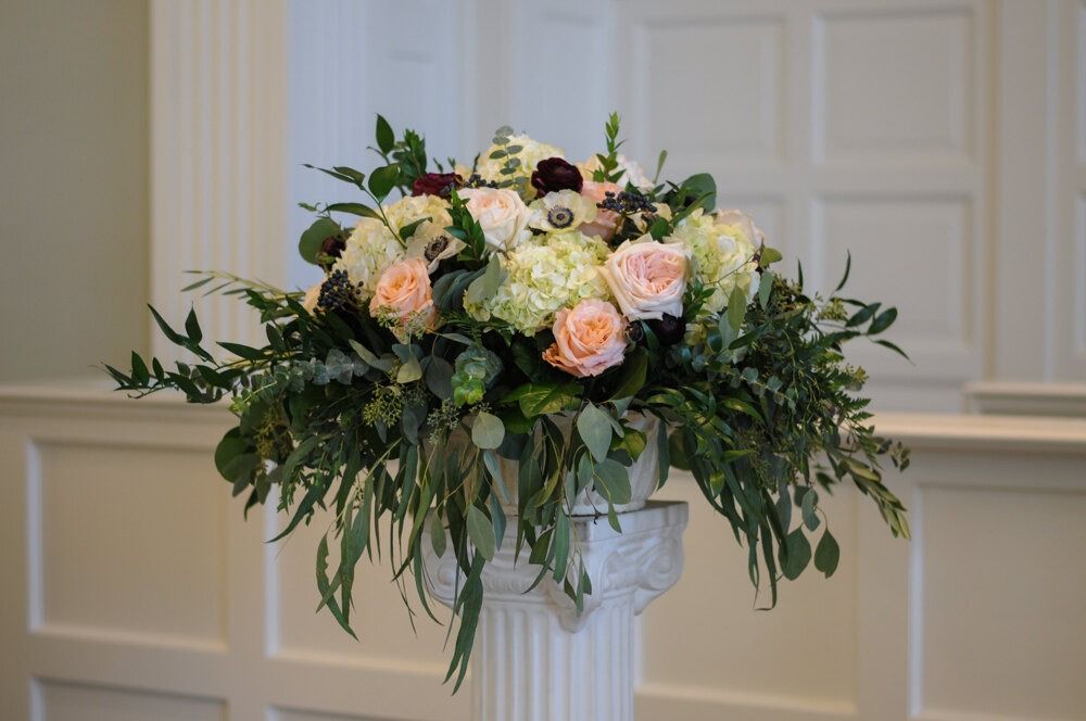 Floral arrangement in white urn on white column; pastel roses, greenery.