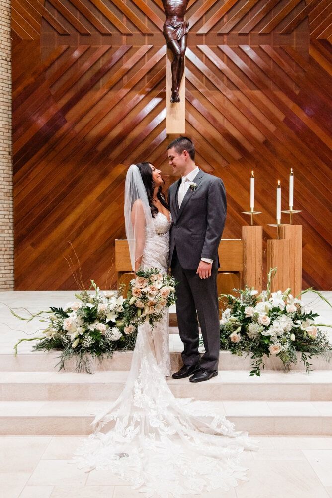 Newlyweds gaze at each other in a church. Bride in white gown with veil; groom in gray suit. Floral arrangements. Wooden backdrop.