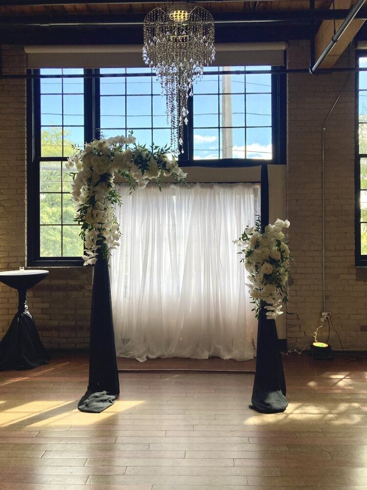 Wedding ceremony setup: floral arch, white curtain backdrop, chandelier, black draping, large windows.