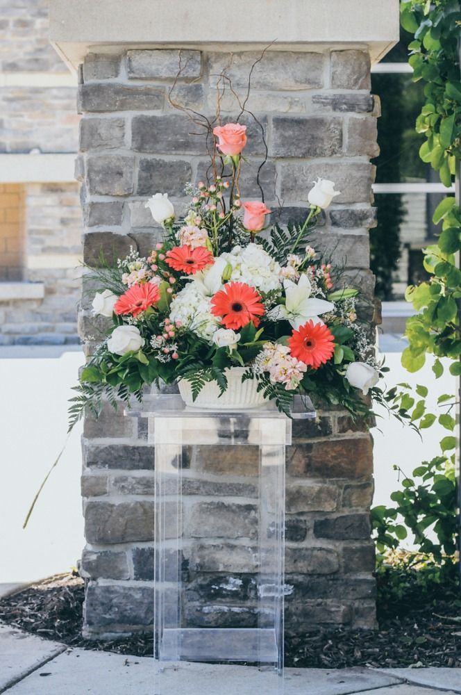 Floral arrangement in white container on clear pedestal, beside brick column.