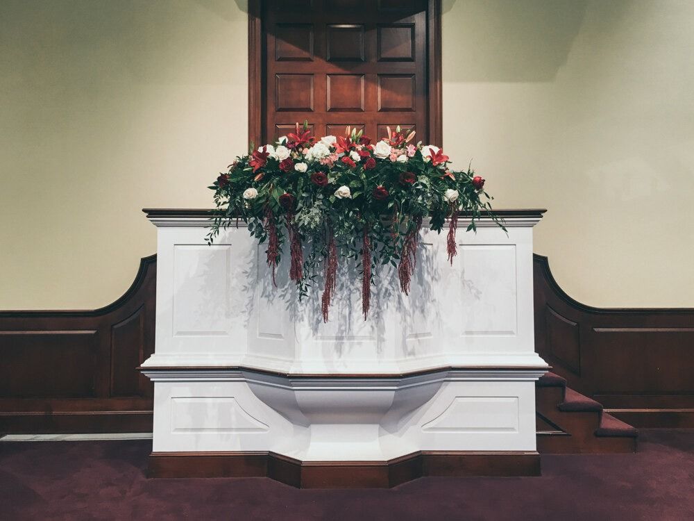 White pulpit with floral arrangement, in a church. Red, white, and green flowers, and brown wooden door background.
