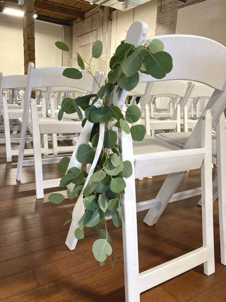 White folding chairs decorated with eucalyptus leaves and ribbon, set up indoors on a wooden floor.