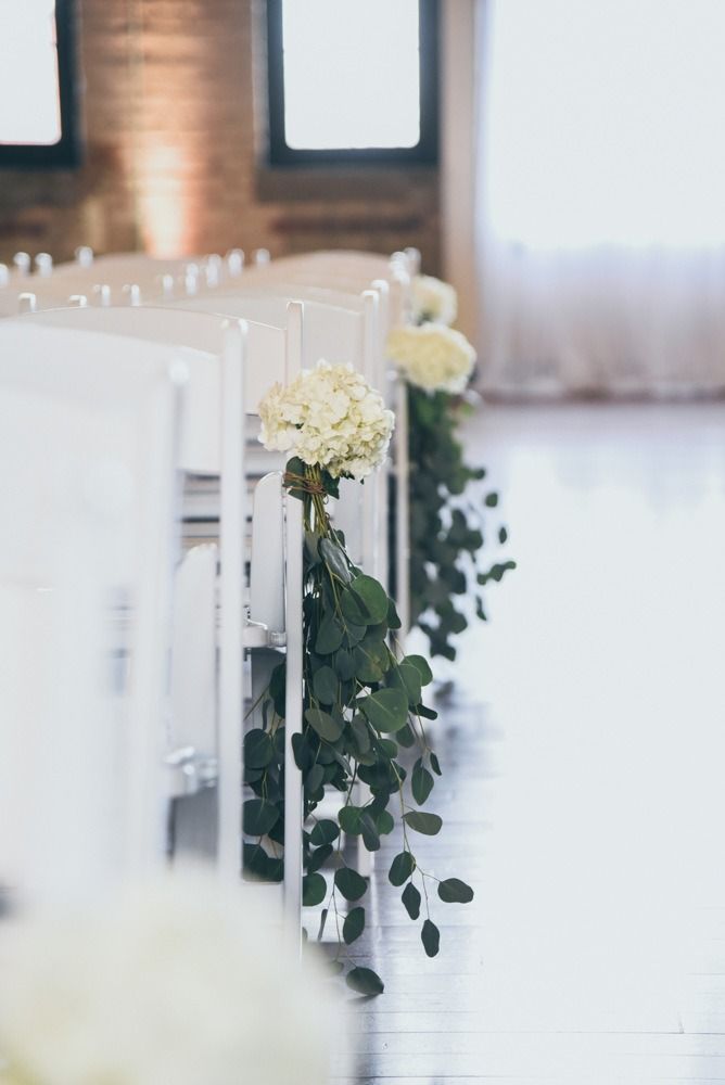 White chairs with floral and greenery decorations line an aisle in a bright room.