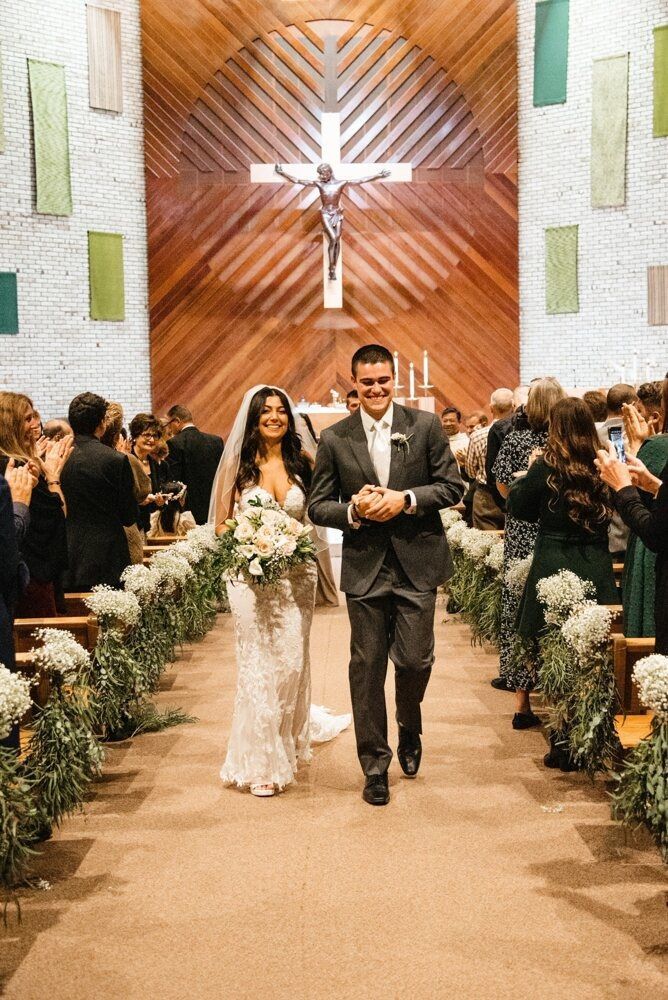 Newlyweds walk down the aisle of a church after their ceremony; guests clap, floral aisle decor.