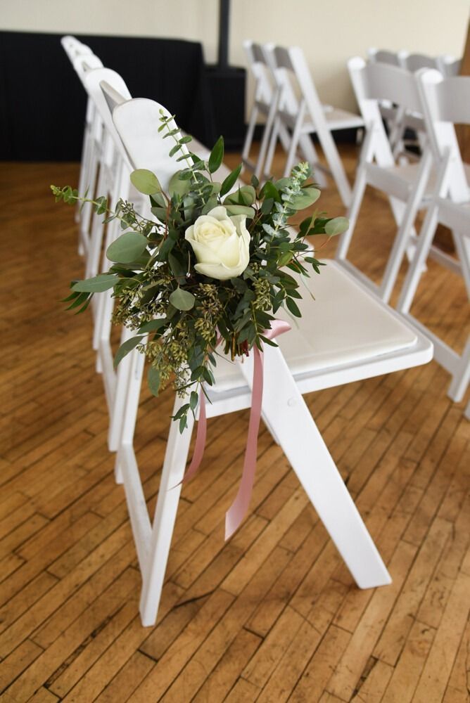 White folding chair decorated with a white rose and greenery, pink ribbon. Wedding setting.