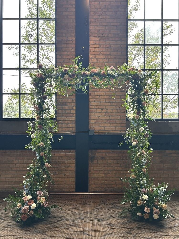 Floral archway with flowers and greenery, in front of a window and brick wall.