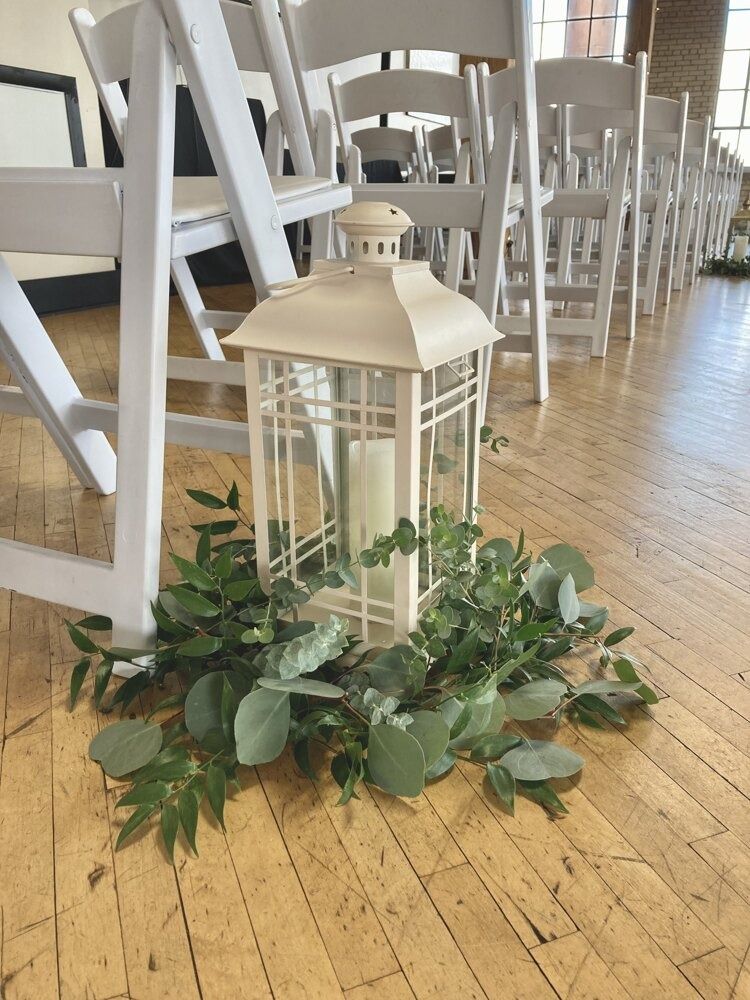 White lantern with candle surrounded by greenery, in front of white chairs, on a wood floor.