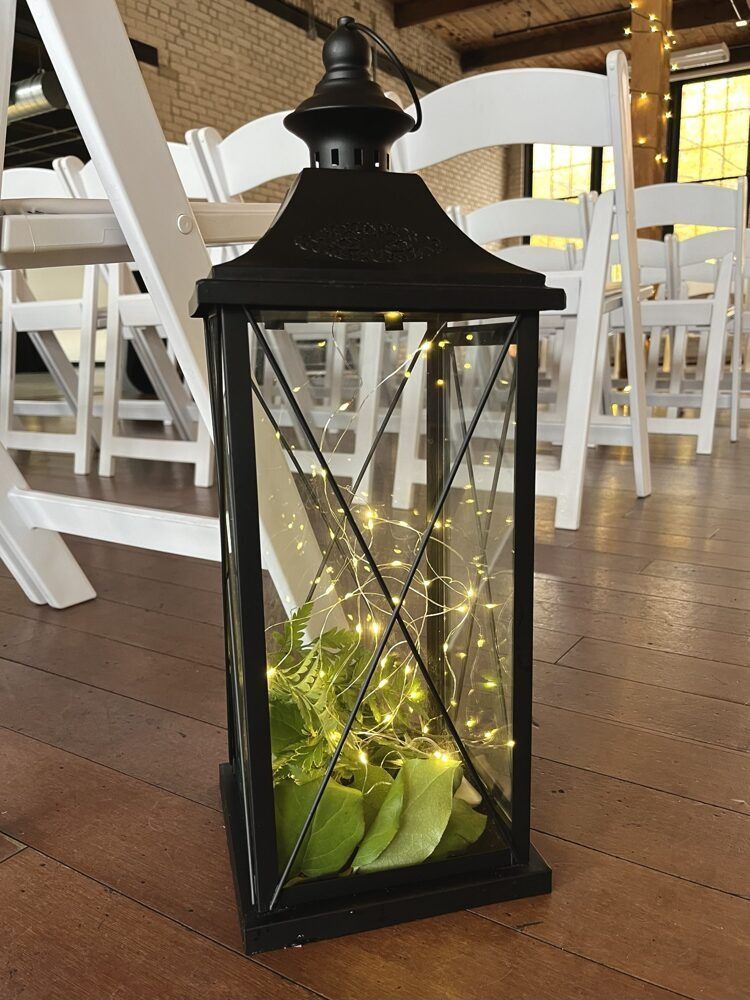 Black lantern with fairy lights and green plants, on a wooden floor, with white chairs in the background.