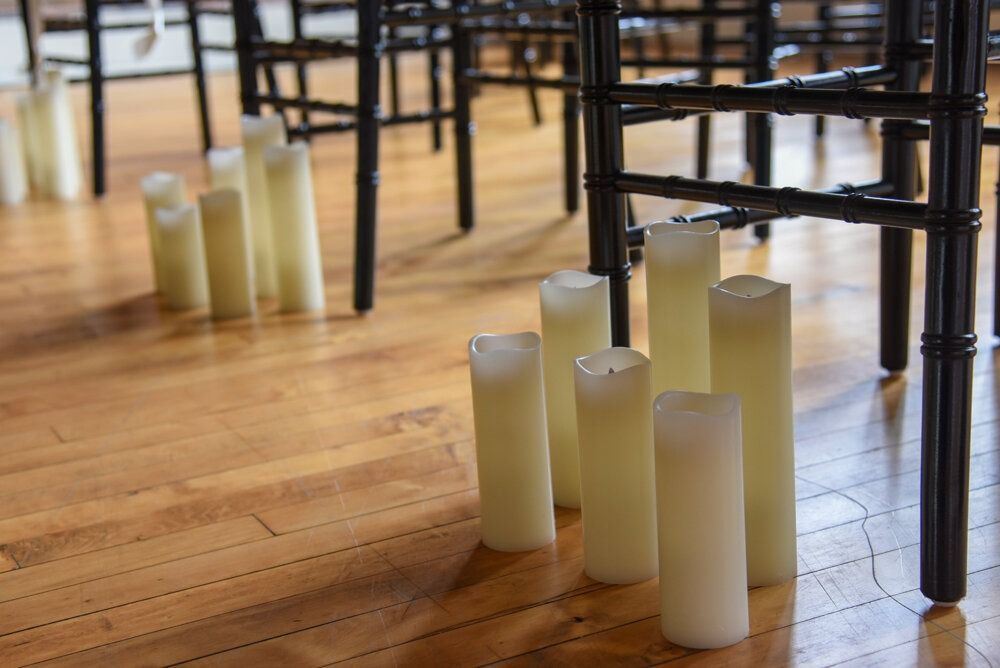 Row of lit white candles on a wooden floor, next to black chairs.