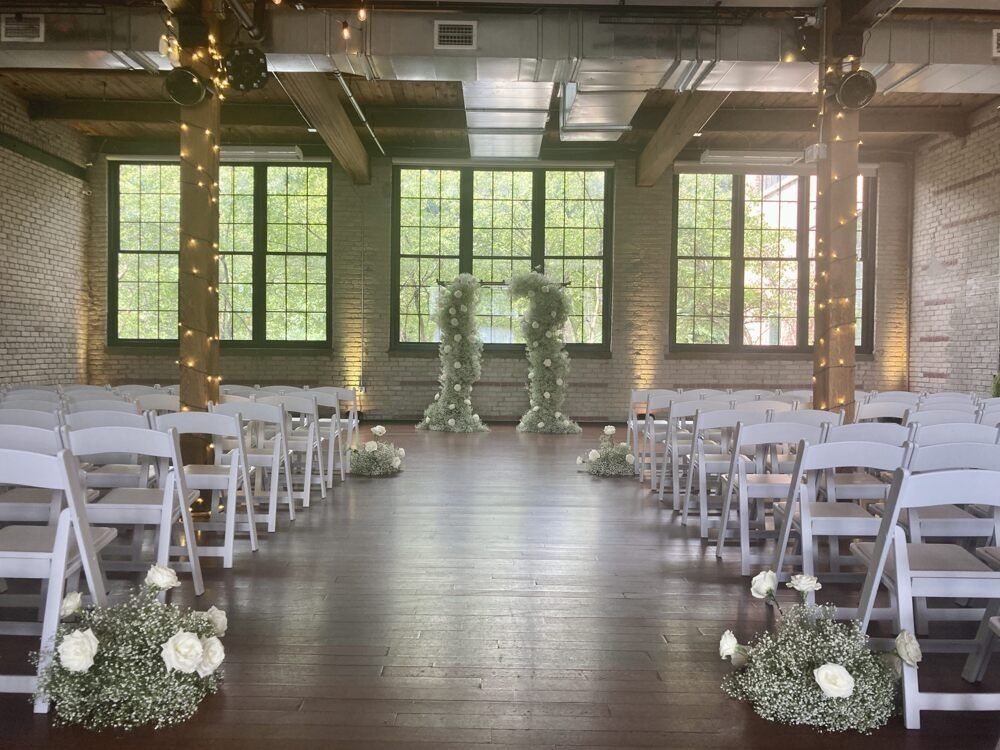 Wedding ceremony setup in a wood-floored room with white chairs, floral arrangements, and archway by windows.
