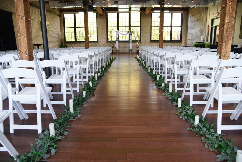 Wedding ceremony setup: white chairs line aisle with greenery, arch at end, indoor venue.
