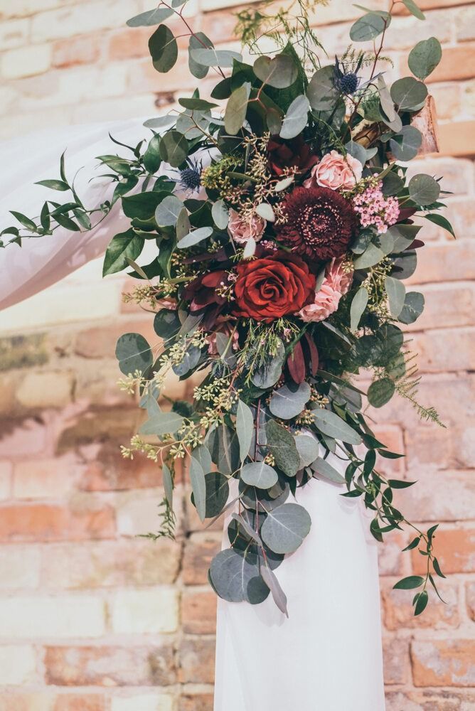 Floral arrangement with red roses and eucalyptus on a white draped structure against a brick wall.