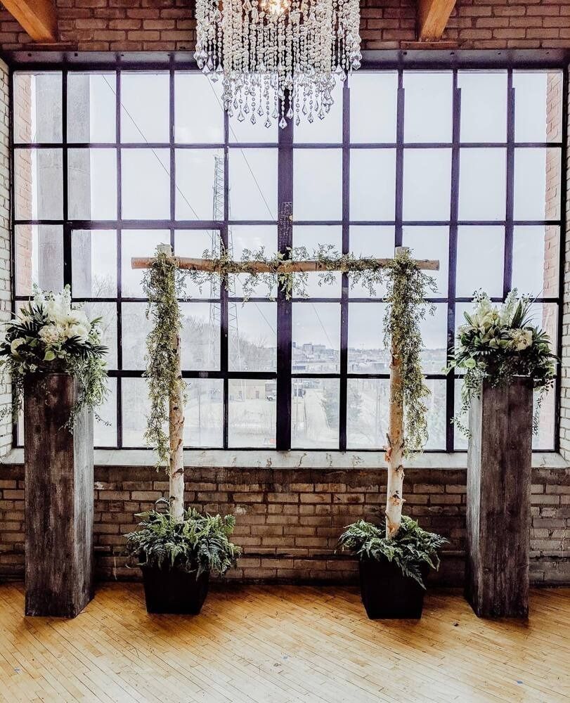 Wedding ceremony setup with archway and floral arrangements in front of a window.