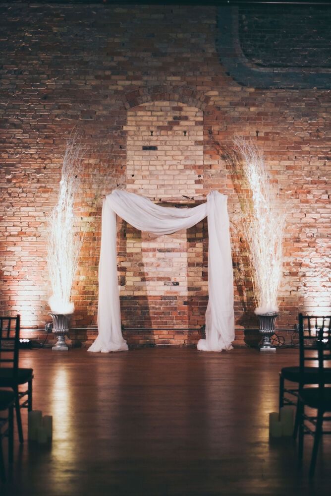 Wedding ceremony setup with draped fabric arch against a brick wall, flanked by lighted branches.