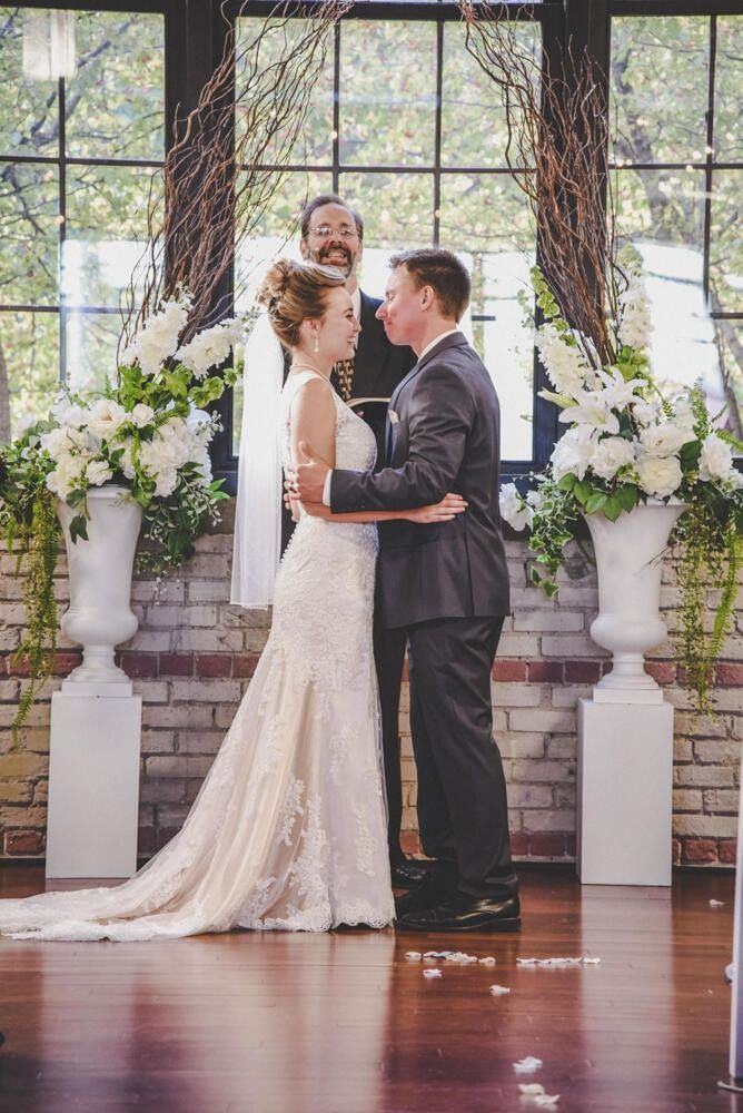 Couple exchanging vows at a wedding ceremony; flowers, brick wall, archway, and officiant in view.