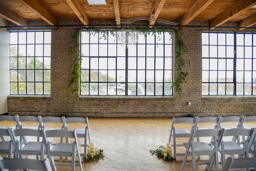 Rows of white chairs face large windows in a venue. Green vines decorate the center window. Wooden beams.