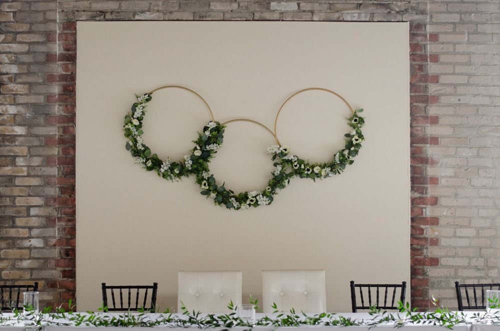 Wedding reception backdrop with floral hoops above a table with chairs, against a brick wall.
