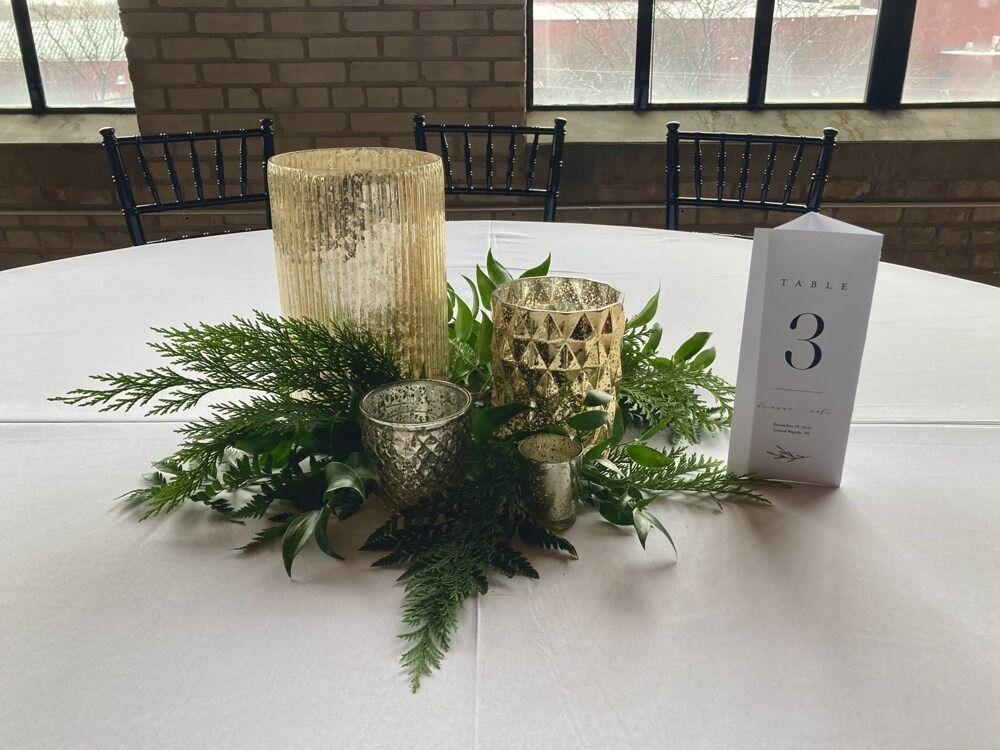 Centerpiece on white tablecloth with candles, greenery, and table number three.