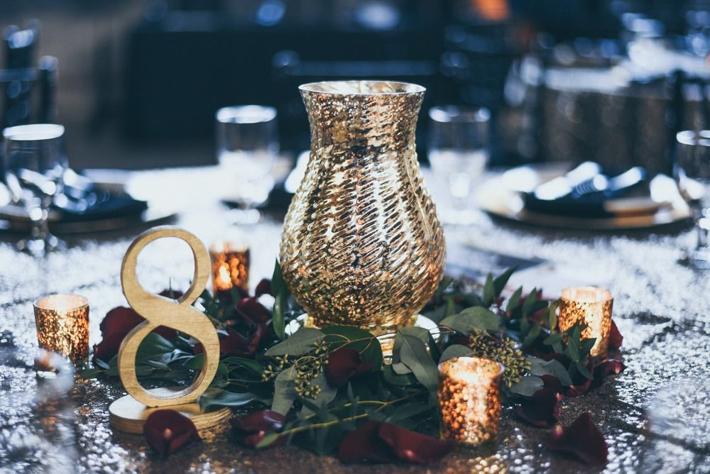Gold vase centerpiece on a table with number 8, candles, red rose petals, and greenery.