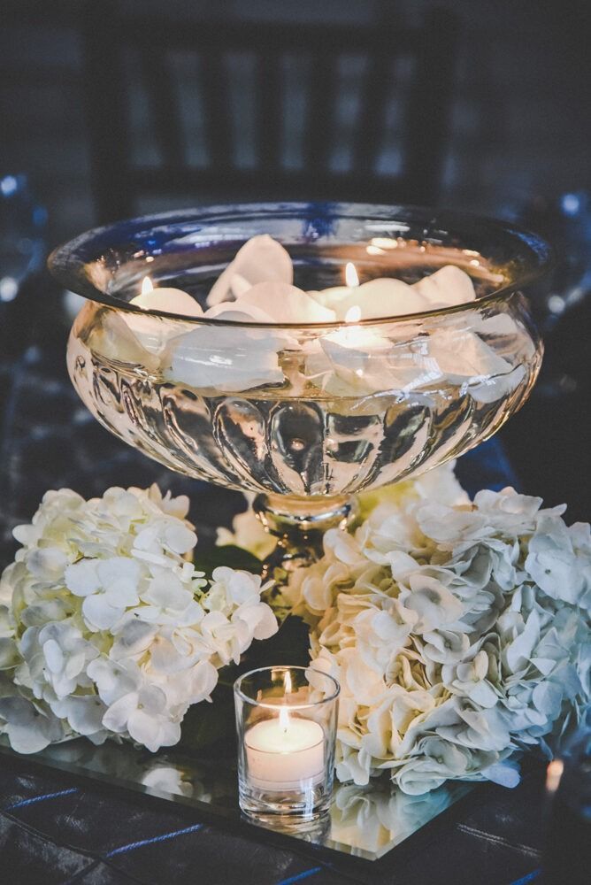 Centerpiece: Clear bowl with floating candles and petals, surrounded by white hydrangeas and a lit votive.