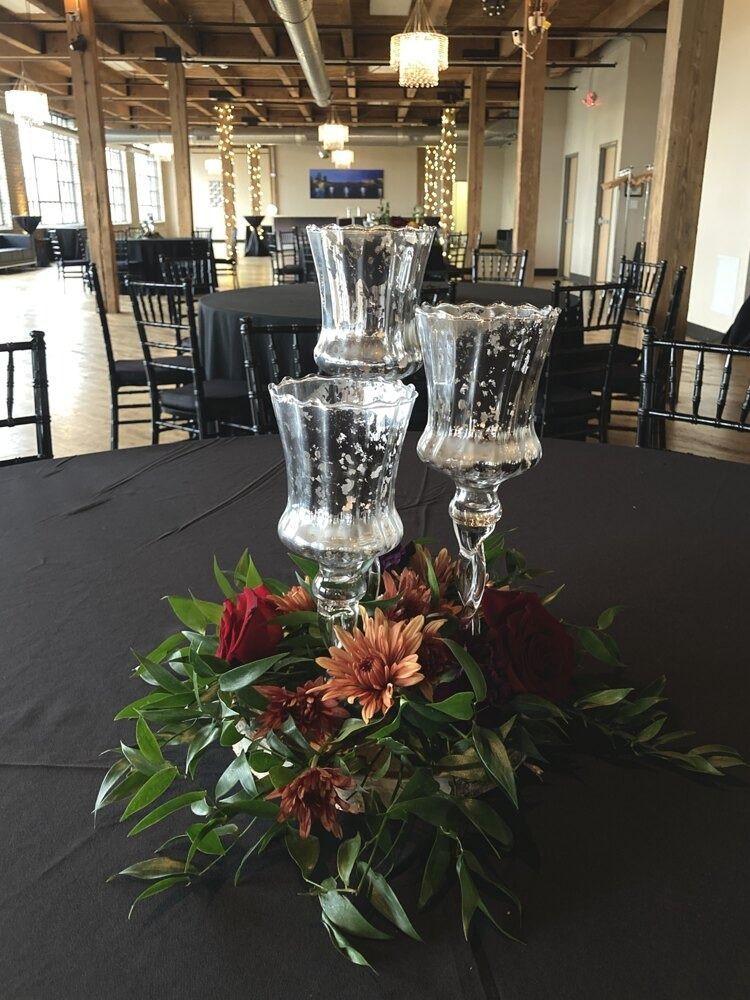 Centerpiece on a black tablecloth: clear glass candle holders atop flowers and greenery. Event setting.