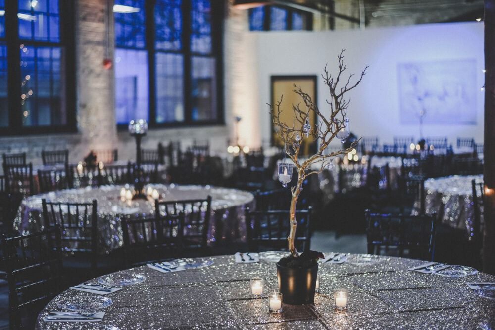 Elegant event space with round tables, shimmering tablecloths, and a decorative tree centerpiece.