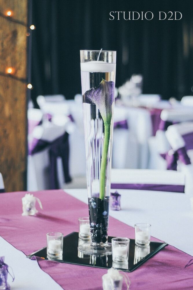 Centerpiece with purple flower inside a tall glass vase filled with water. Mirror base, candles, purple table runner.