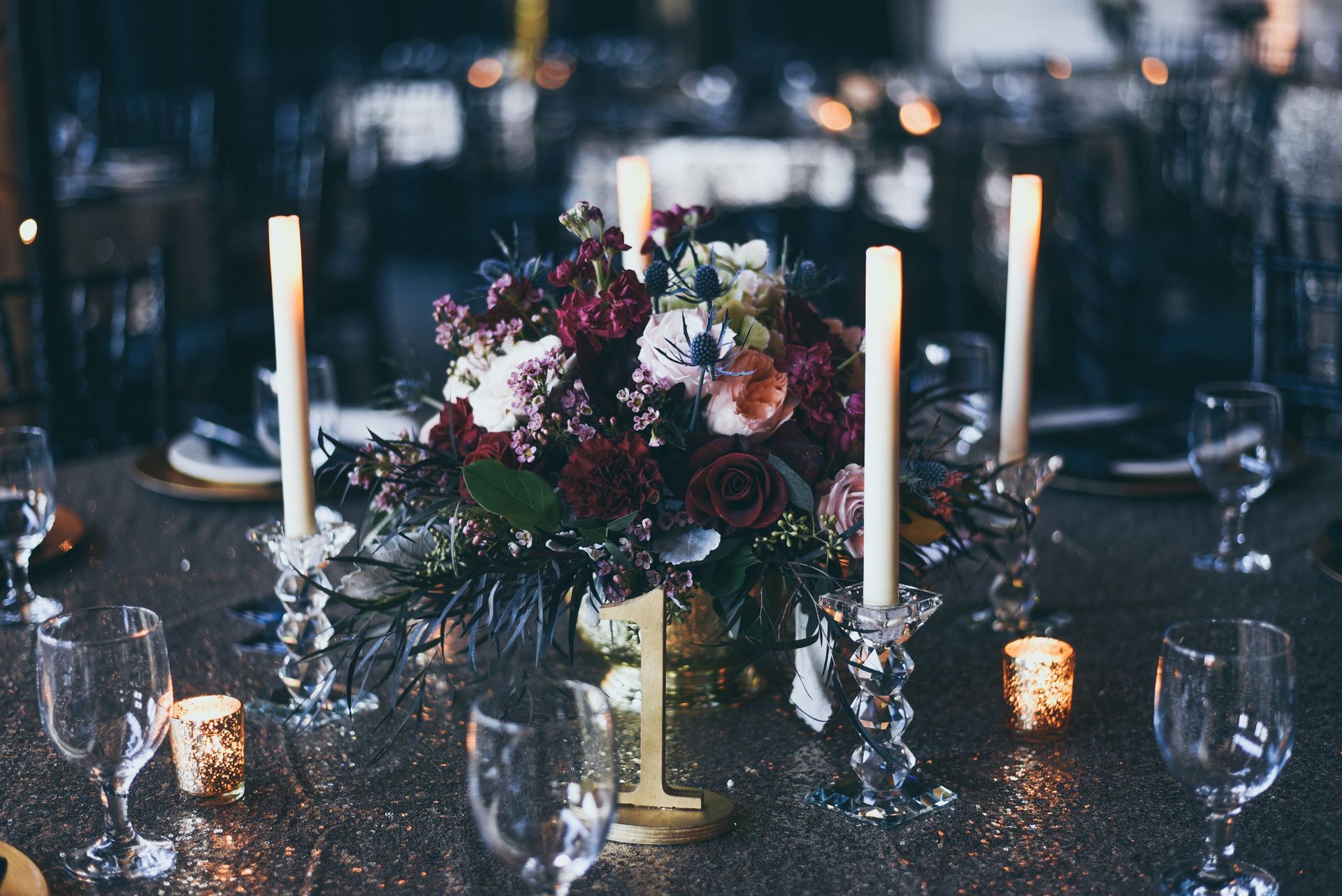 Centerpiece with flowers, candles, and table number on a golden tablecloth in a dimly lit setting.
