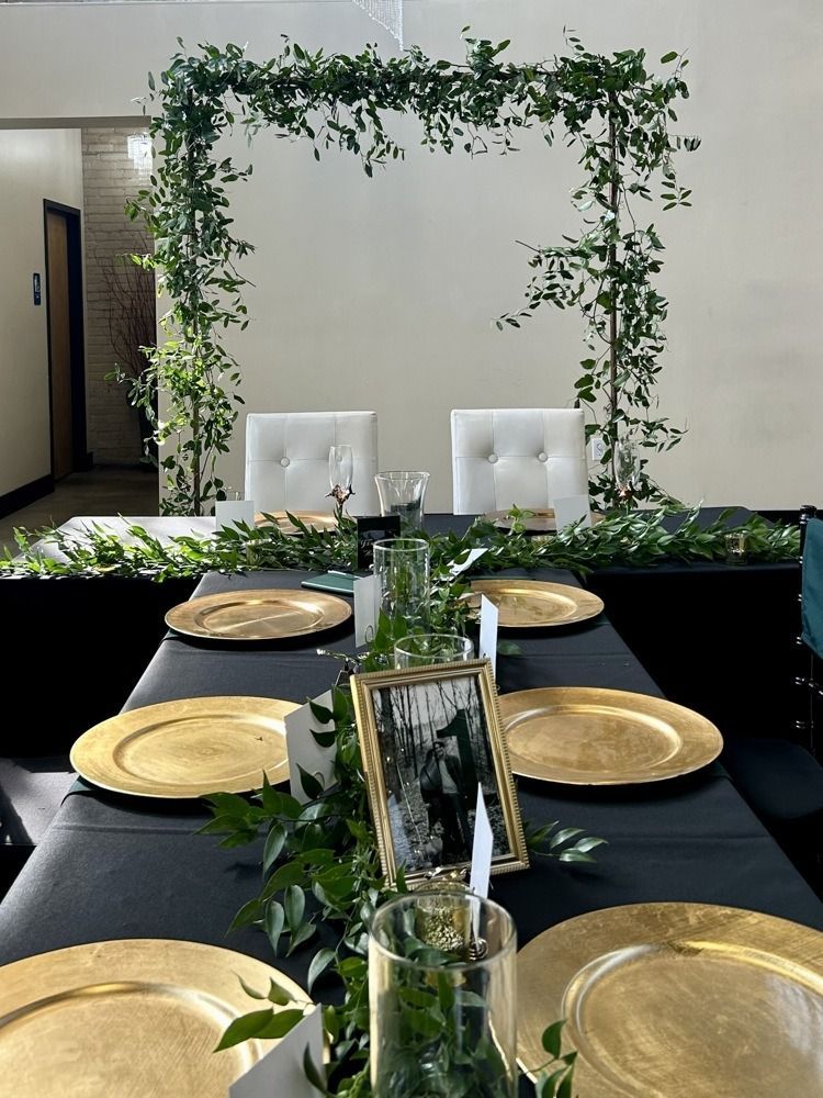 Wedding reception table with gold chargers, greenery, and arch decorated with vines.