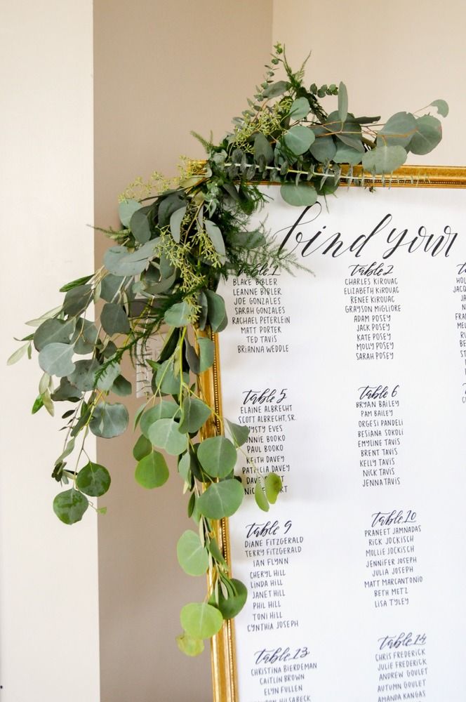 Wedding seating chart with greenery, gold frame on white surface, guests' names listed.