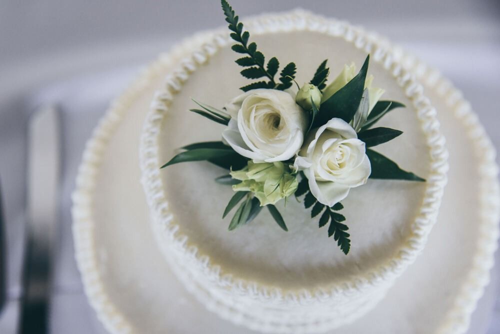 White tiered cake decorated with white roses and greenery on a white plate.