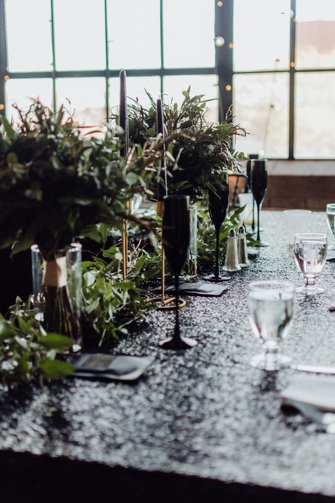 Elegant table setting with black champagne flutes, greenery, and a sparkling black tablecloth in front of a window.