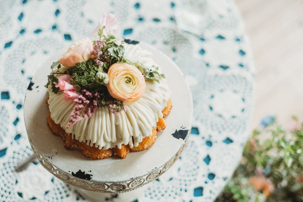 Cake with white frosting and flowers on a decorative stand, set on a lacy tablecloth.