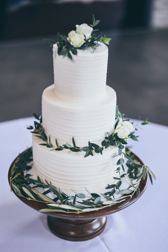 Three-tiered white wedding cake with floral and green leaf accents on a wooden cake stand.