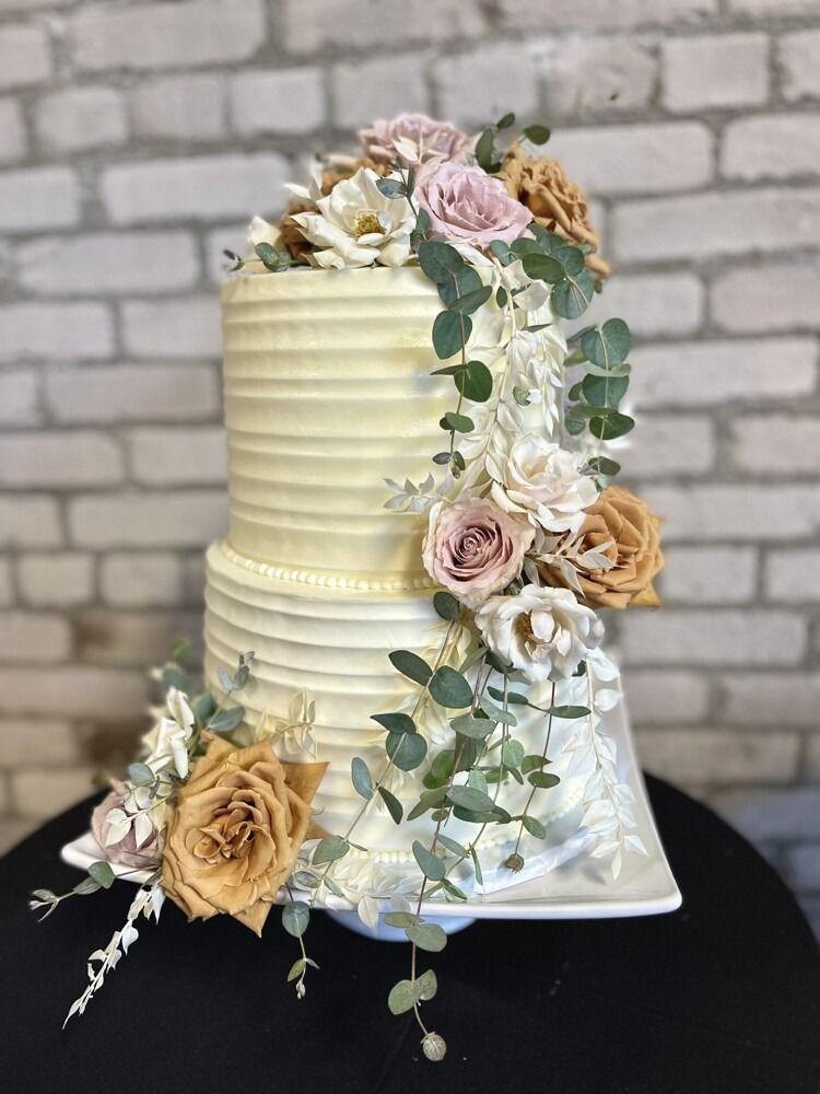 Two-tiered wedding cake with textured buttercream frosting, floral accents, and greenery against a brick wall.