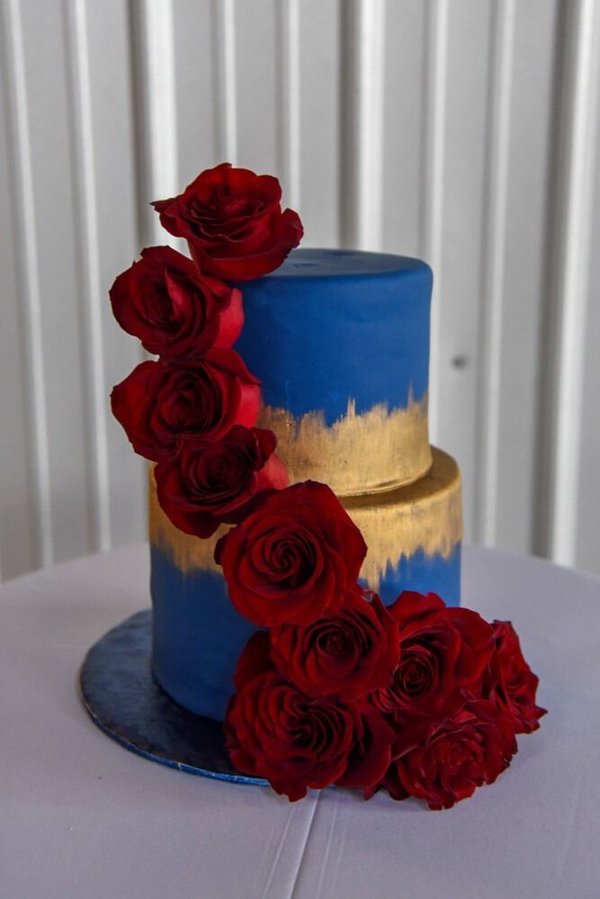 Two-tiered blue cake with gold brushstrokes, adorned with red roses on a white tablecloth.