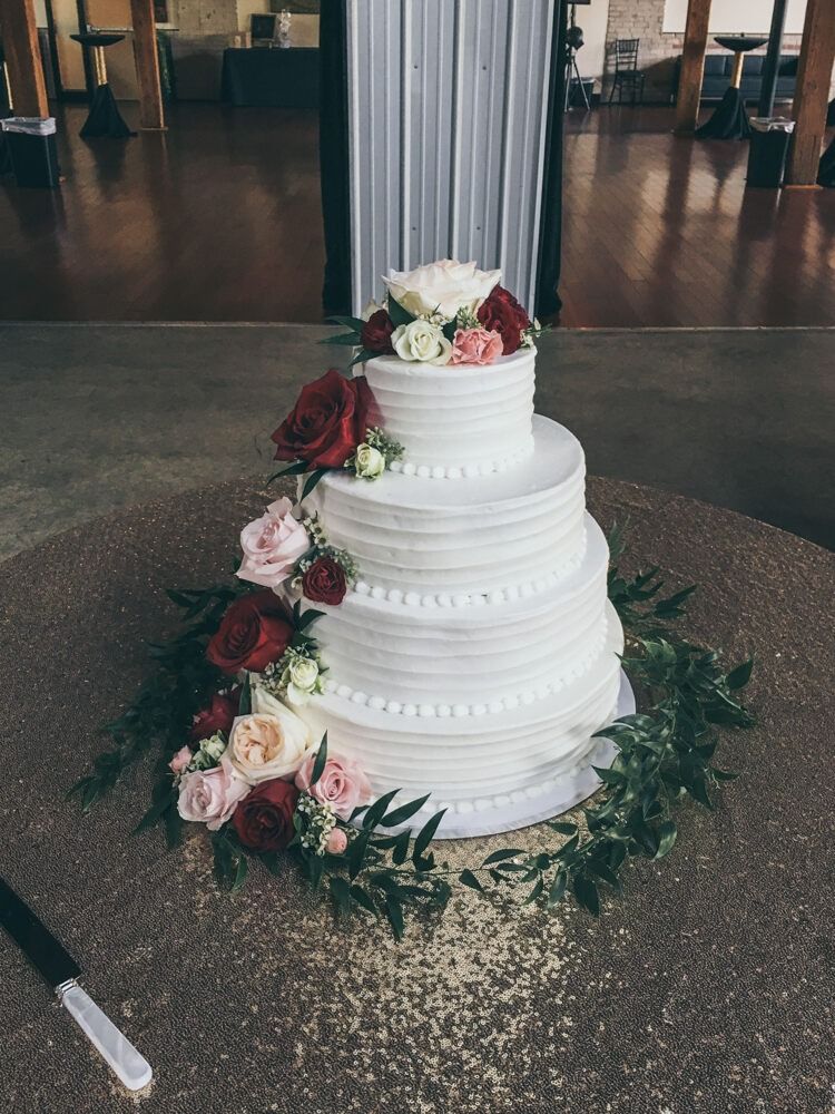 Four-tier white wedding cake decorated with red and pink roses and greenery on a gold sequin tablecloth.