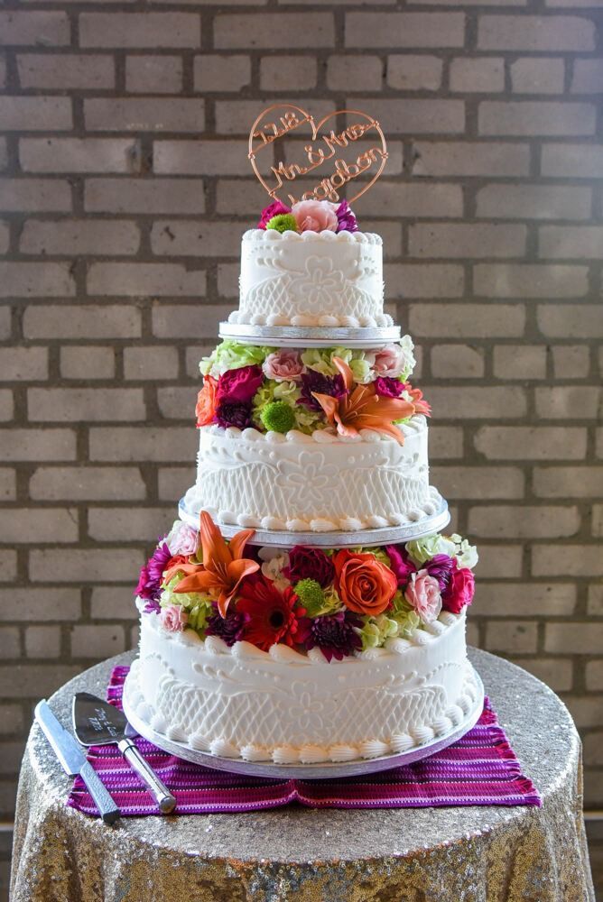 Five-tiered wedding cake with white frosting, decorated with flowers and a heart cake topper, on a sequined table.
