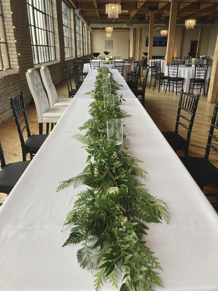 Long table set for an event, decorated with greenery and candles in a brightly lit room.