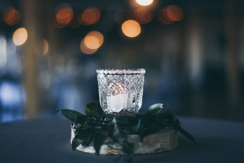 Candle in decorative glass holder on a wood base, with greenery, and blurred lights in background.