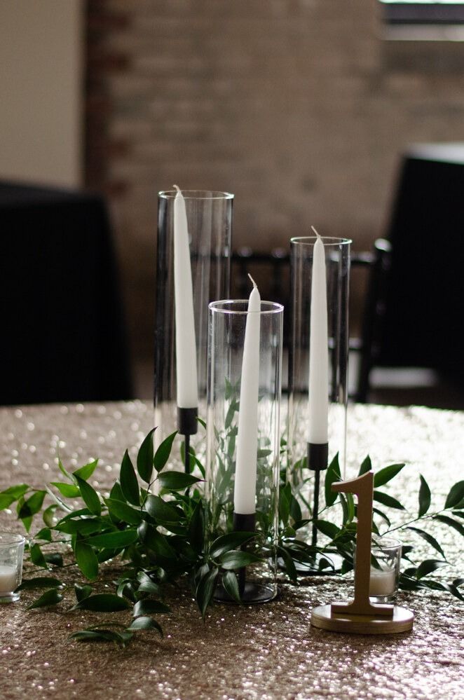 Table centerpiece with gold sequin tablecloth, glass cylinder vases holding white taper candles, and greenery with a gold table number.