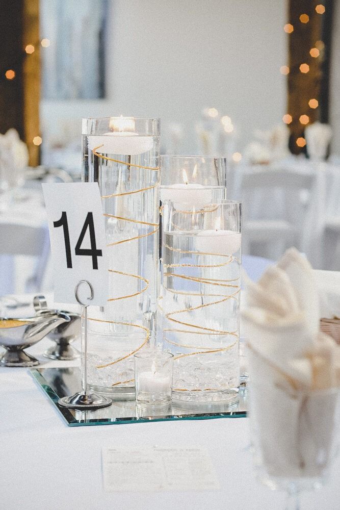 Centerpiece on a table at a wedding. Cylindrical vases hold water, floating candles, and gold string, on a mirrored surface.