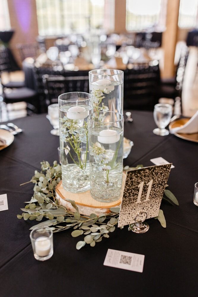 Wedding table centerpiece with three glass cylinders, floating candles, and greenery on a wooden base. Table number 11.