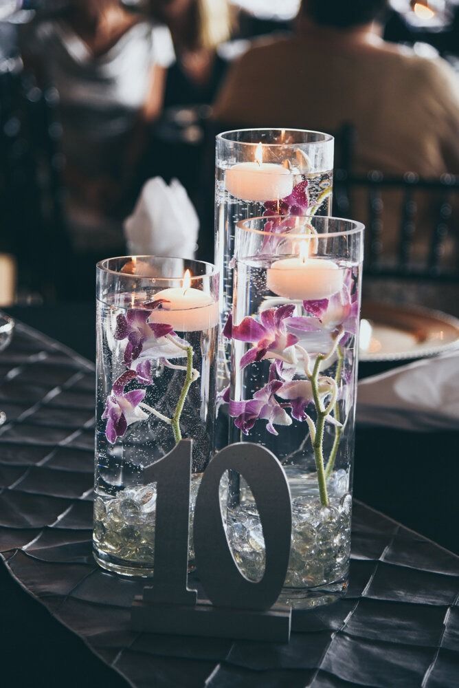 Table centerpiece with three glass vases filled with water, orchids, and floating candles; table number 10.