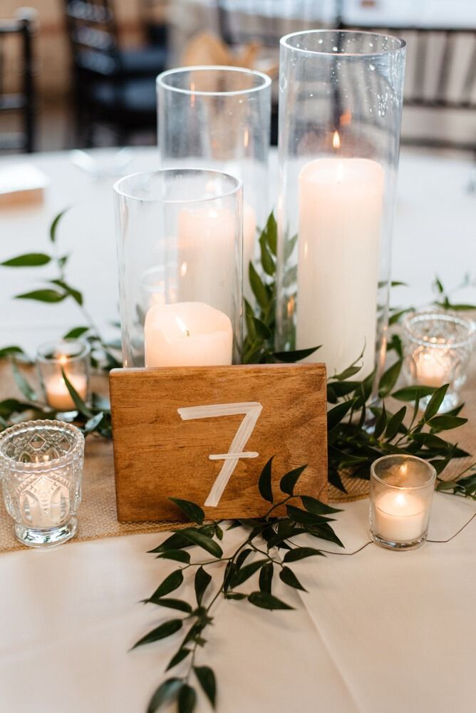 Wedding table centerpiece: candles in glass cylinders, wooden table number 7, greenery, and small votive candles.