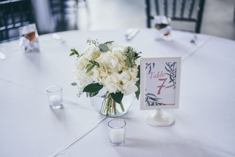 White floral centerpiece and table number 7 on a white tablecloth; two candles and a glass.