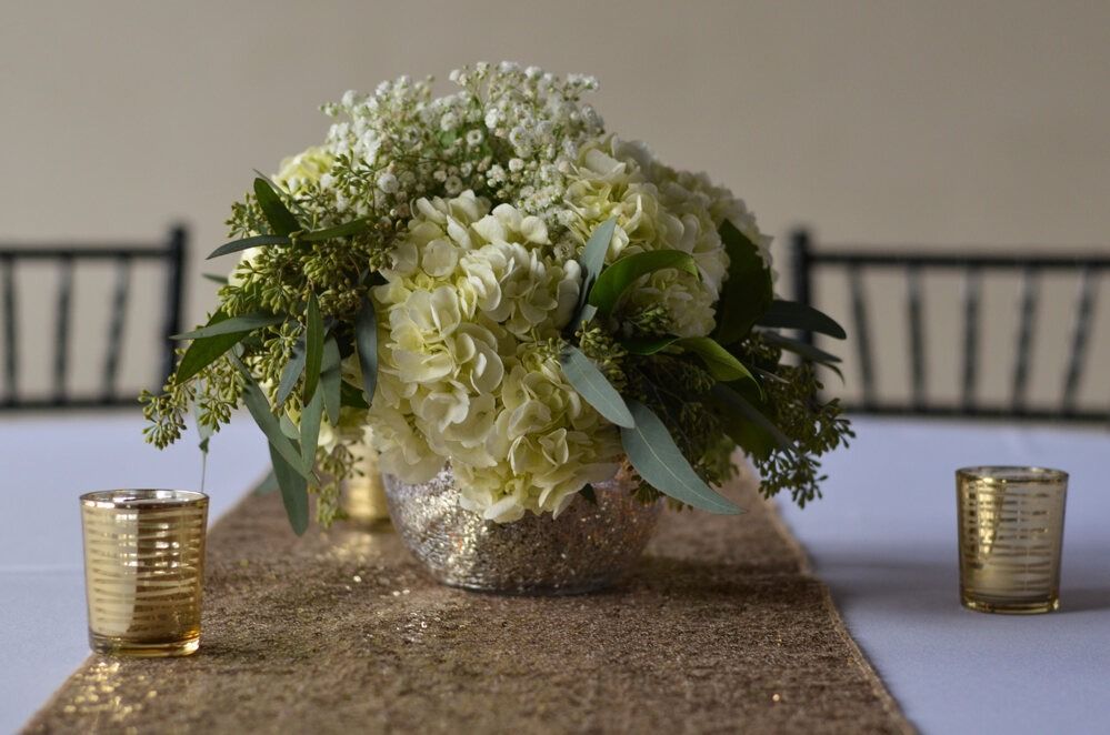 Centerpiece of white flowers, greenery, and gold accents on a table with candles.