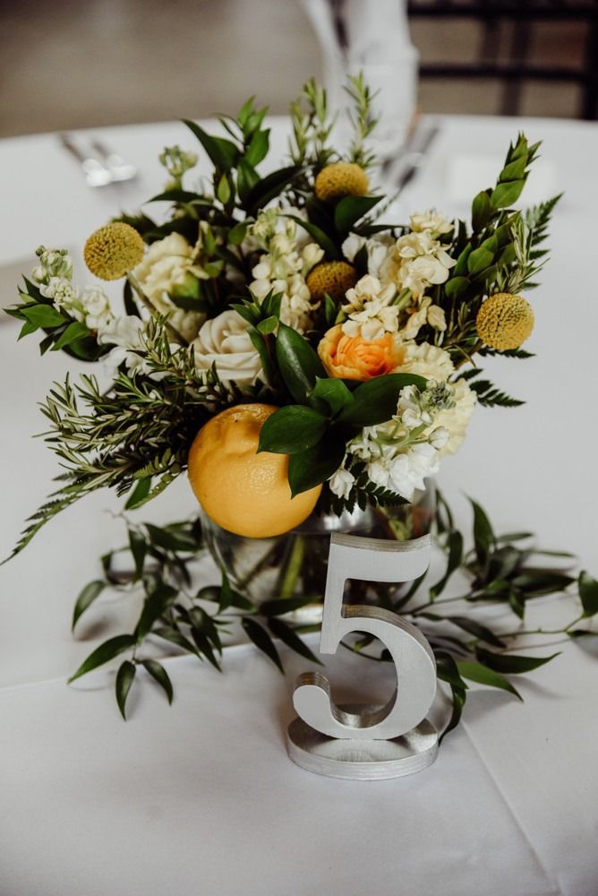 Centerpiece with orange, white flowers, greenery, and table number five on a white tablecloth.