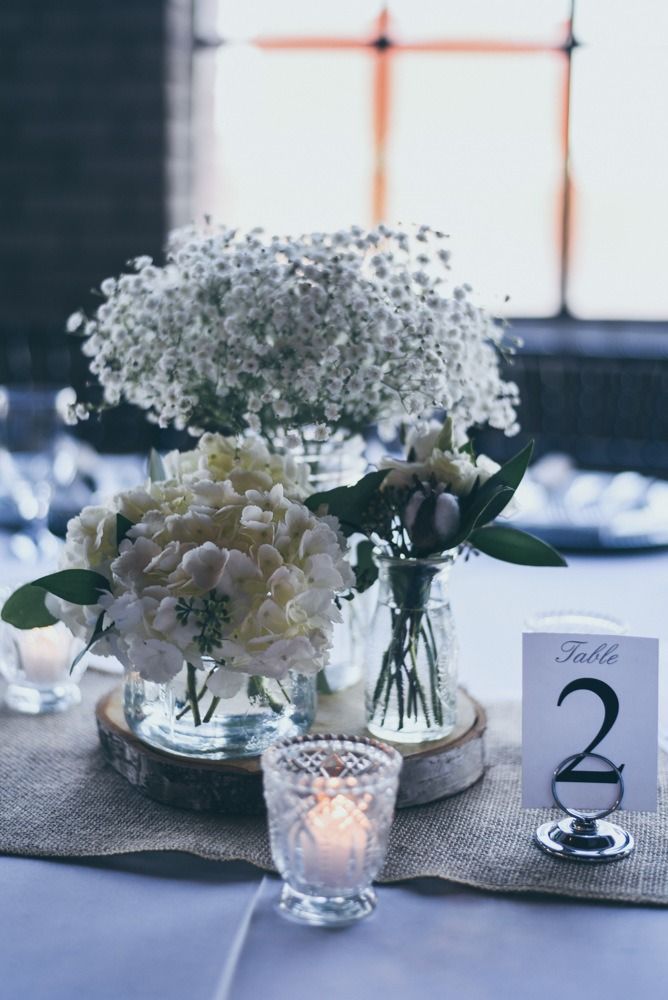Centerpiece with white flowers in glass jars on a wooden slice and a table number, on a burlap runner.